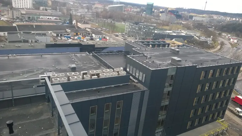Plokščias daugiaaukščio pastato stogas su bitumine danga, parapetais ir vėdinimo įranga, miestas fone